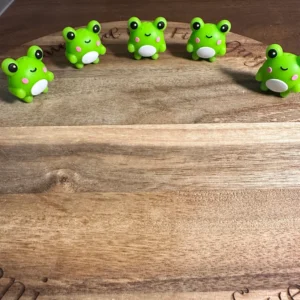 Four 3D frog silicone focal beads with white bellies and pink cheeks are lined up on a wooden surface.