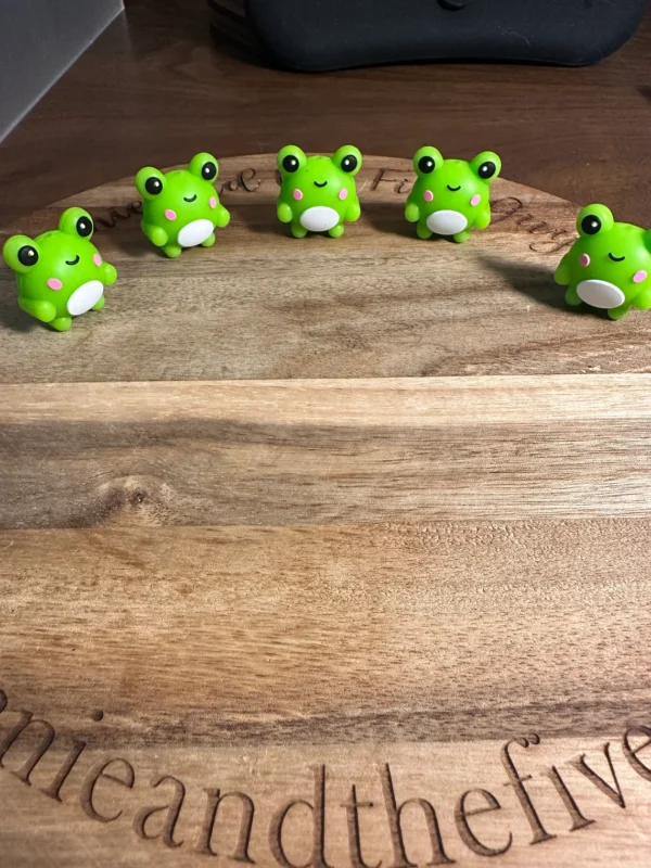 Four 3D frog silicone focal beads with white bellies and pink cheeks are lined up on a wooden surface.