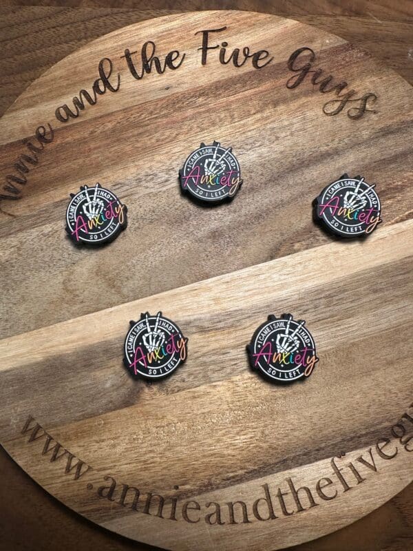 Five commemorative pins with "celebrating 10 years" and "AnnieLy" text displayed on a wooden board with "Annie and the Five Guys" and "www.annieandthefiveguys.com" text around the edge are replaced by Your anxiety is a lying hoe silicone focal bead.