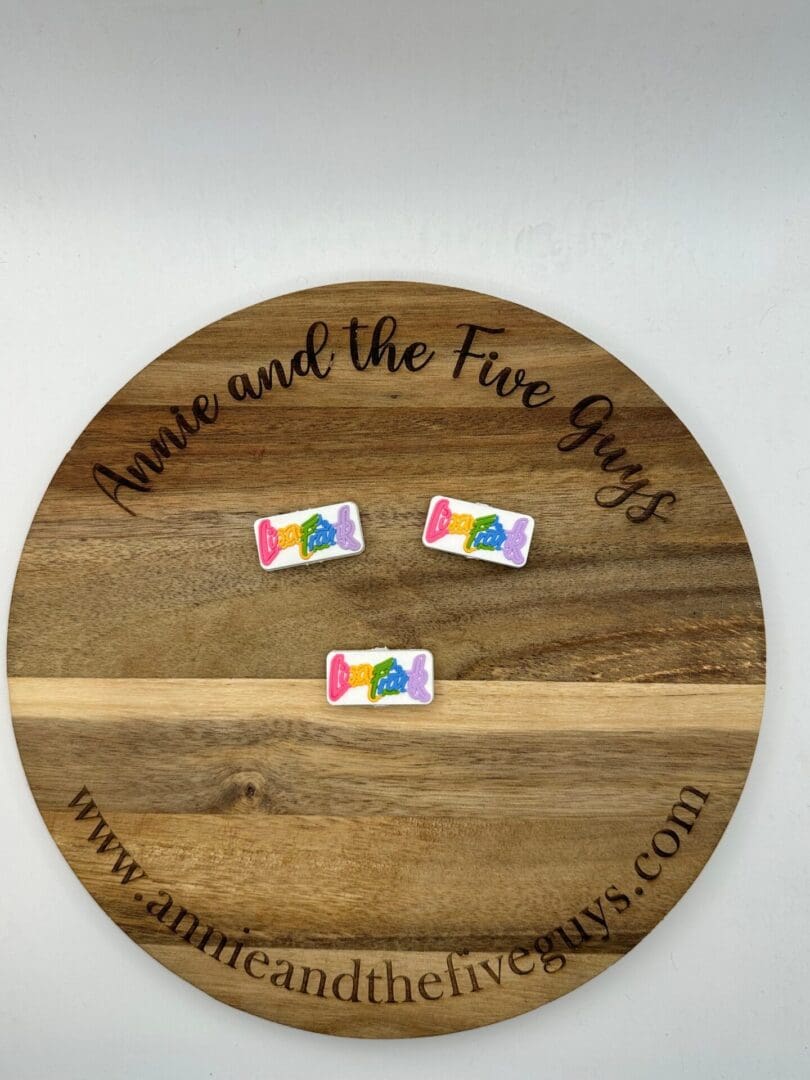 Wooden round board with the text "Annie and the Five Guys" and three "LF silicone focal bead" colorful candies on it. Website link "www.annieandthefiveguys.com" is also visible at the bottom of the board.