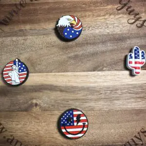 A circular wooden board displays four American-themed pins: an eagle on a US flag patriotic silicone focal bead, the Statue of Liberty with a flag background, a cactus with a flag design, and a flag heart with a bolt.