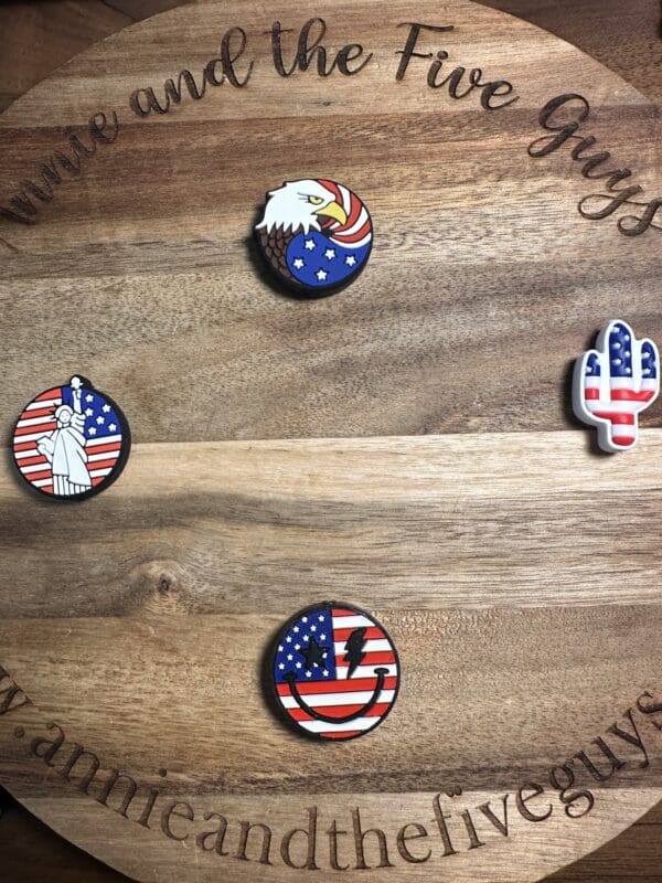 A circular wooden board displays four American-themed pins: an eagle on a US flag patriotic silicone focal bead, the Statue of Liberty with a flag background, a cactus with a flag design, and a flag heart with a bolt.