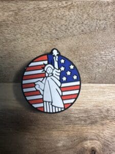 A round enamel pin, named "Eagle on US flag patriotic silicone focal bead," features a white Statue of Liberty set against the red and white stripes and blue field with stars of the American flag. It rests on a wooden surface.