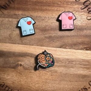 Displayed on a wooden surface with text around the edge, you will find two enamel pins shaped like medical scrubs with hearts—one in blue and one in pink—alongside a pin featuring a cheetah cuddling a stethoscope.