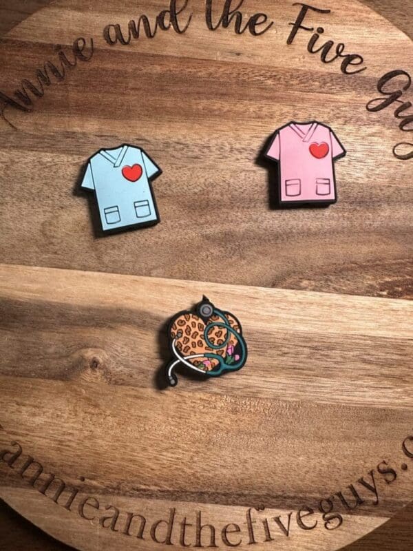 Displayed on a wooden surface with text around the edge, you will find two enamel pins shaped like medical scrubs with hearts—one in blue and one in pink—alongside a pin featuring a cheetah cuddling a stethoscope.