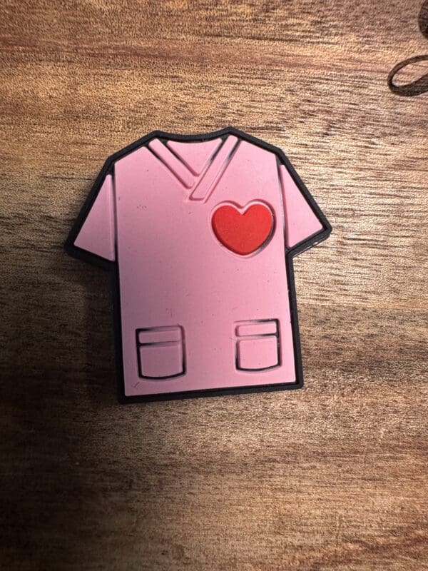 A Stethoscope silicone focal bead designed as a pink hospital scrub top with a red heart on the chest, lying on a wooden surface.