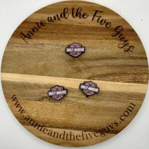 A round wooden board featuring the text "Annie and the Five Guys" with a motorcycle focal bead placed on it. The website "www.annieandthefiveguys.com" is also visible on the board.