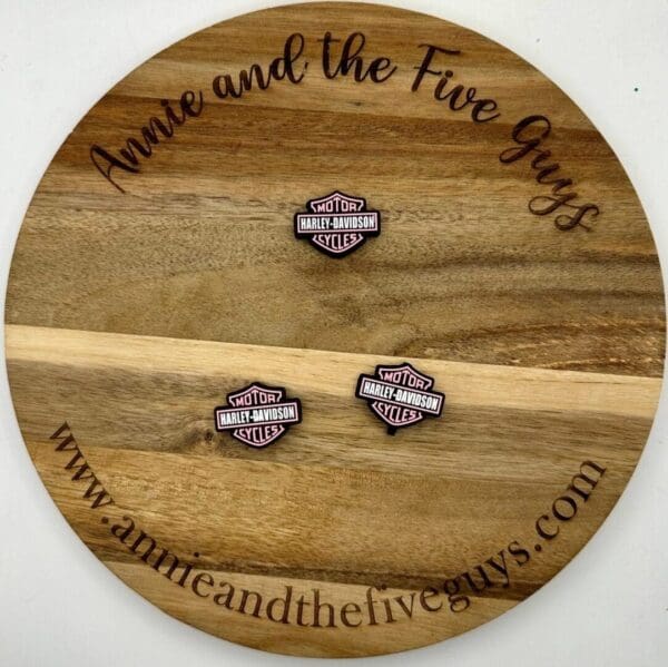 A round wooden board featuring the text "Annie and the Five Guys" with a motorcycle focal bead placed on it. The website "www.annieandthefiveguys.com" is also visible on the board.