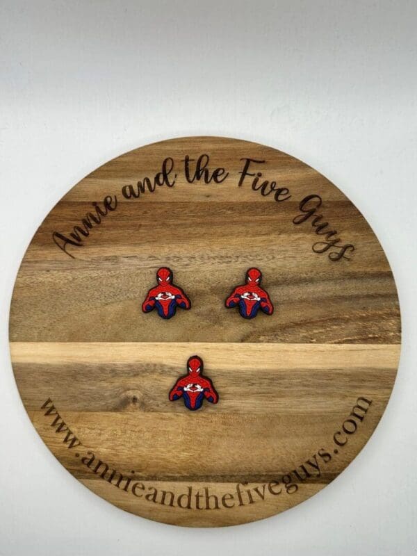 A round wooden board with the text "Annie and the Five Guys" features three small Spider guy focal beads. The website "www.annieandthefiveguys.com" is also printed on the board's rim.