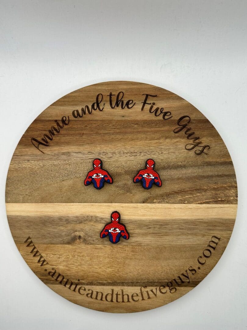 A round wooden board with the text "Annie and the Five Guys" features three small Spider guy focal beads. The website "www.annieandthefiveguys.com" is also printed on the board's rim.