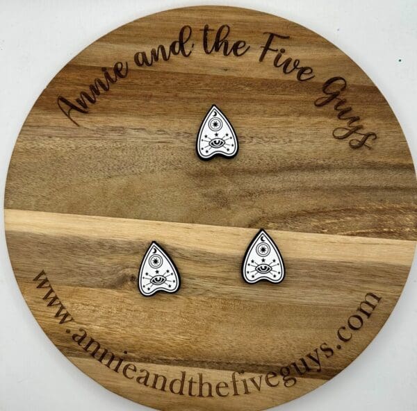 Wooden round board with "Annie and the Five Guys" written on the edge. Three small, white, teardrop-shaped Planchette silicone focal bead items with a black-outlined design are placed on the board. Web address is visible.