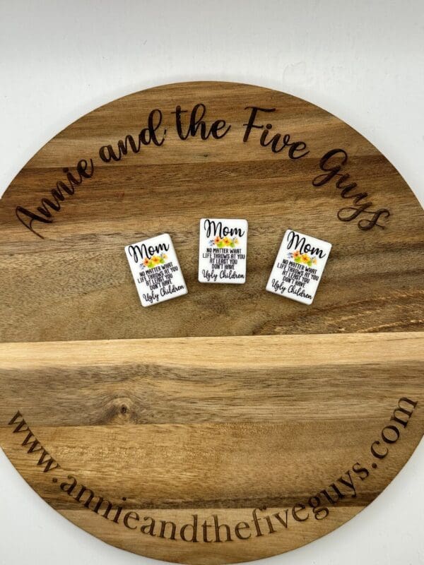 A round wooden board with the words "Annie and the Five Guys" and the website "www.annieandthefiveguys.com," displaying three small Mom silicone focal beads.