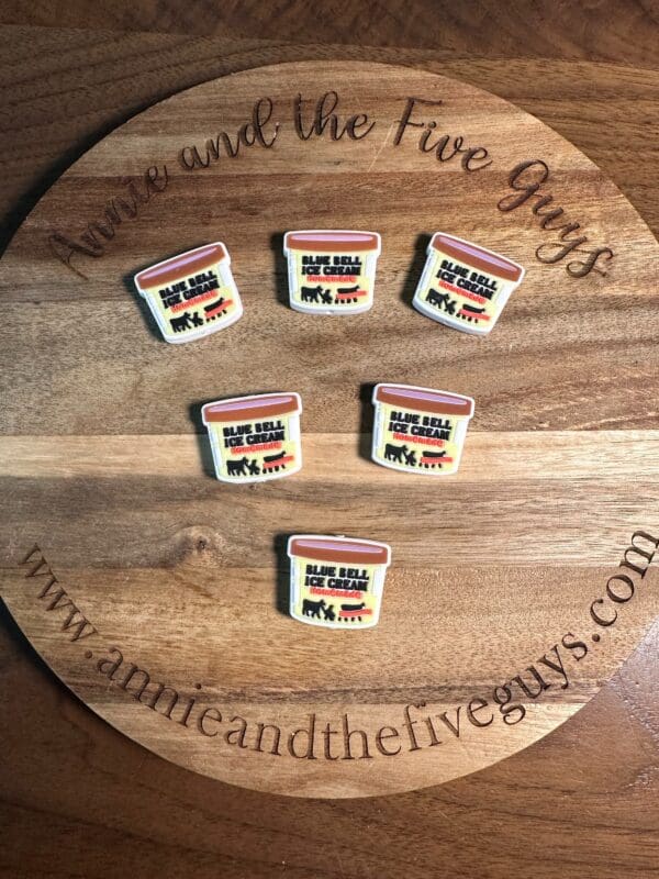 Five small pins resembling Ice cream silicone focal beads are placed on a round wooden surface with the text “Annie and the Five Guys” and a website address.