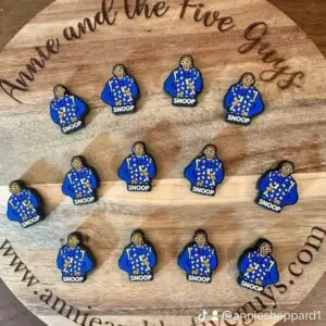 A circular wooden board displays 14 identical custom collab silicone focal beads featuring a cartoon character in a blue outfit. The text around the board reads "Annie and the Five Guys" and includes a social media handle.