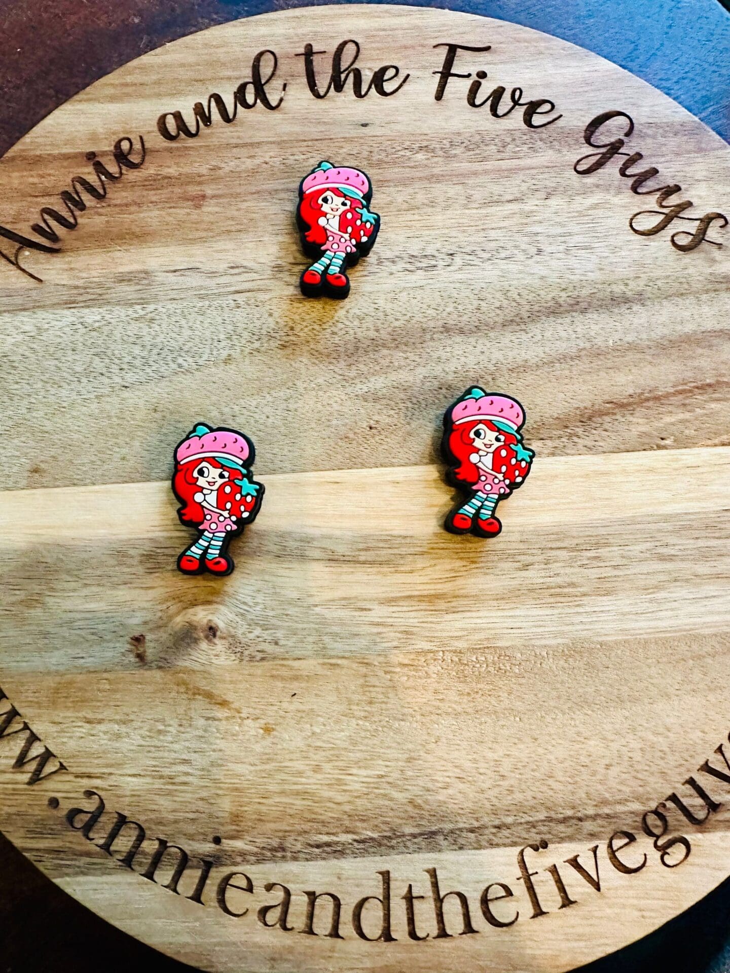 Three cartoon character pins placed on a round wooden board with the text "Annie and the Five Guys" engraved in a circular pattern.