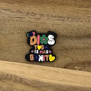 A colorful pin on a wooden surface reads "Con Dios, todo es mas bonito" with flowers and hearts around the text.