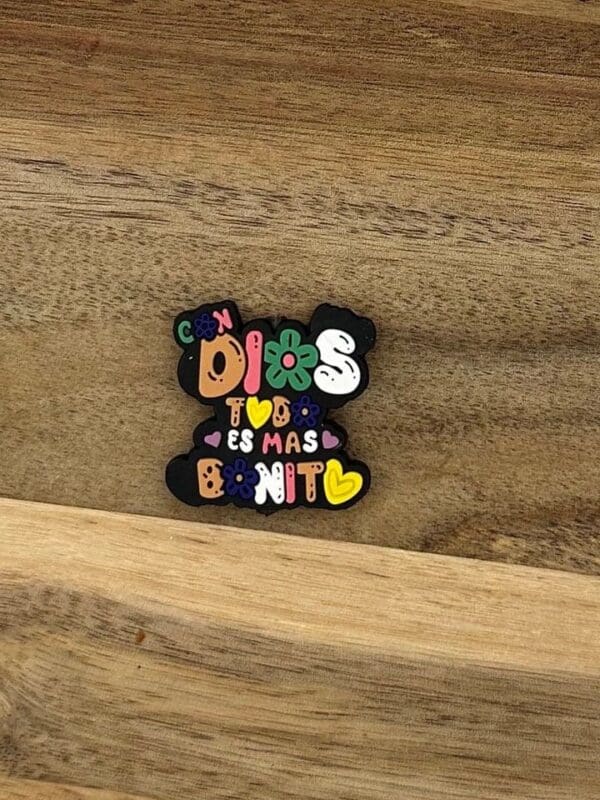 A colorful pin on a wooden surface reads "Con Dios, todo es mas bonito" with flowers and hearts around the text.