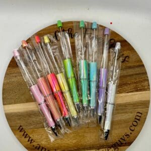 A set of Opalescent Beadable pens in various pastel colors, each individually wrapped in plastic, displayed on a wooden board.