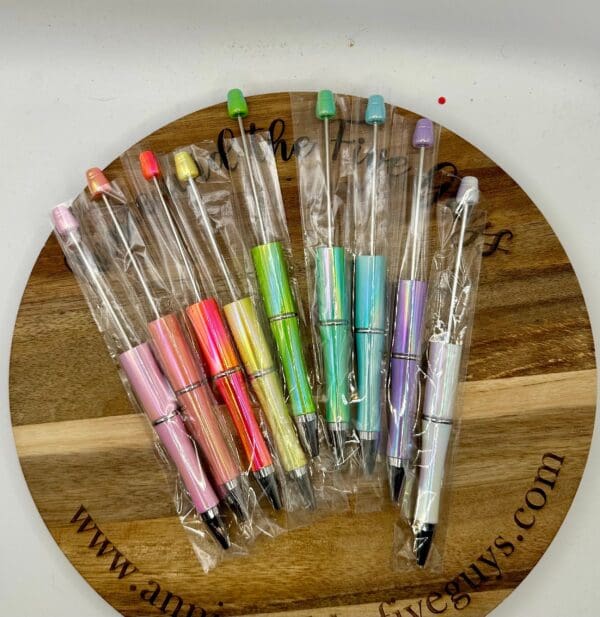 A set of Opalescent Beadable pens in various pastel colors, each individually wrapped in plastic, displayed on a wooden board.