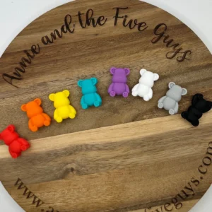 Seven small Bear silicone focal beads in various colors (red, orange, yellow, teal, purple, white, gray, and black) are arranged in a row on a round wooden board.