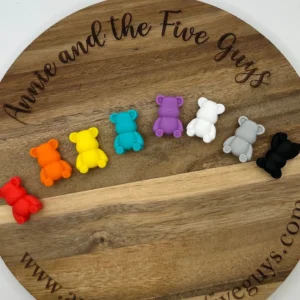 Seven small Bear silicone focal beads in various colors (red, orange, yellow, teal, purple, white, gray, and black) are arranged in a row on a round wooden board.