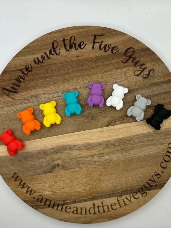 Seven small Bear silicone focal beads in various colors (red, orange, yellow, teal, purple, white, gray, and black) are arranged in a row on a round wooden board.