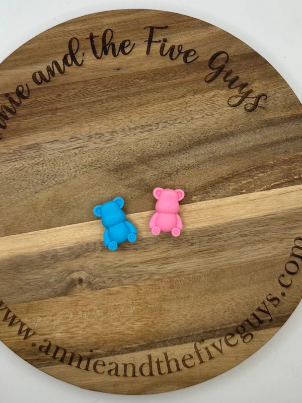 Two small Bear silicone focal beads, one blue and one pink, placed on a round wooden board with the text "Annie and the Five Guys" engraved around the edge.