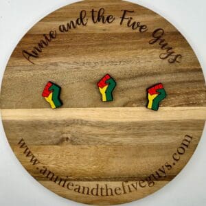 A round wooden board featuring three fist-shaped pins, colored in green, yellow, and red. The board is engraved with the text "Annie and the Five Guys" and the website "www.annieandthefiveguys.com".