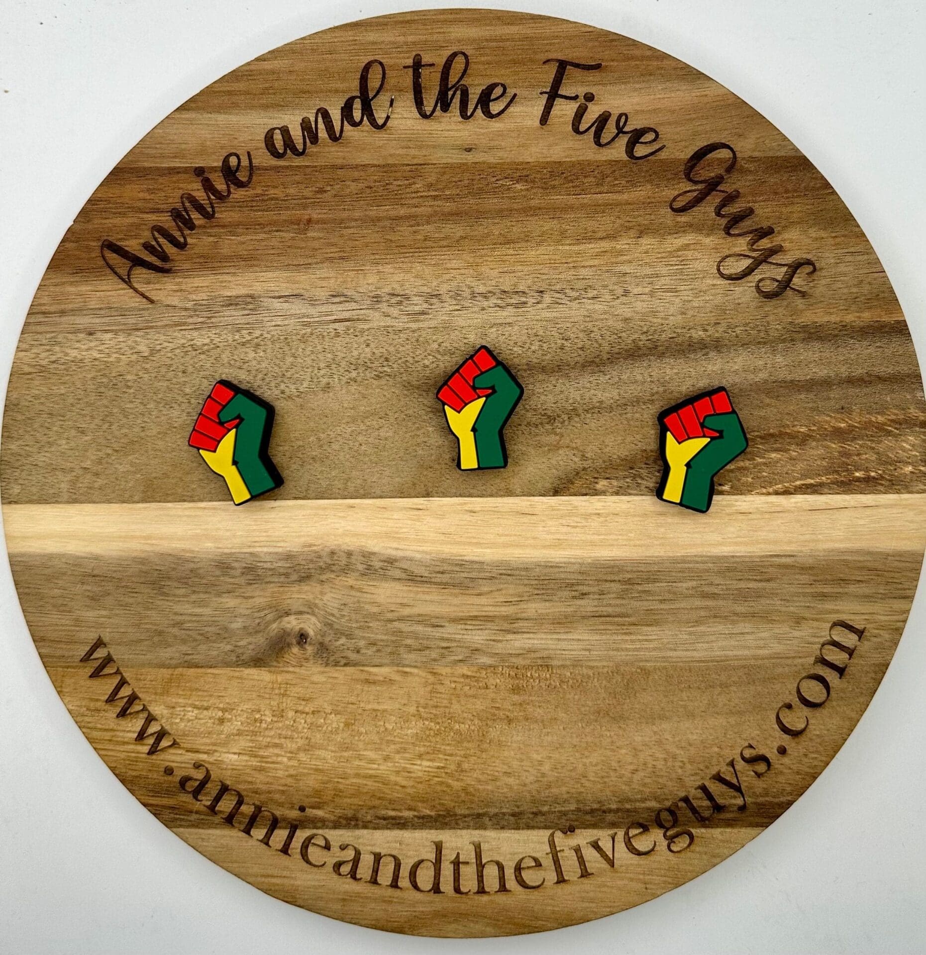 A round wooden board featuring three fist-shaped pins, colored in green, yellow, and red. The board is engraved with the text "Annie and the Five Guys" and the website "www.annieandthefiveguys.com".