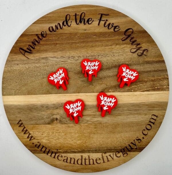 A circular wooden board with "Annie and the Five Guys" inscribed on the edge displays five red and white "Baby Bunny" pins. Website www.annieandthefiveguys.com is printed at the bottom.