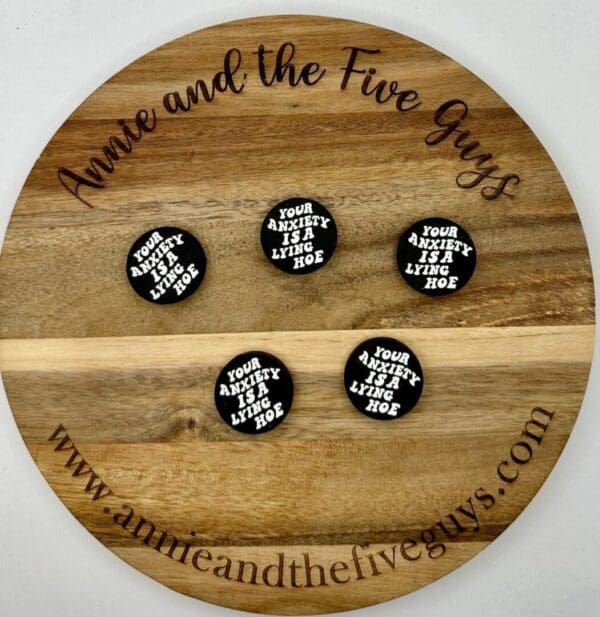 A wooden tray with "Annie and the Five Guys" engraved around the edge displays five round badges that read "Your anxiety is a lying hoe." The website "www.annieandthefiveguys.com" is also engraved.