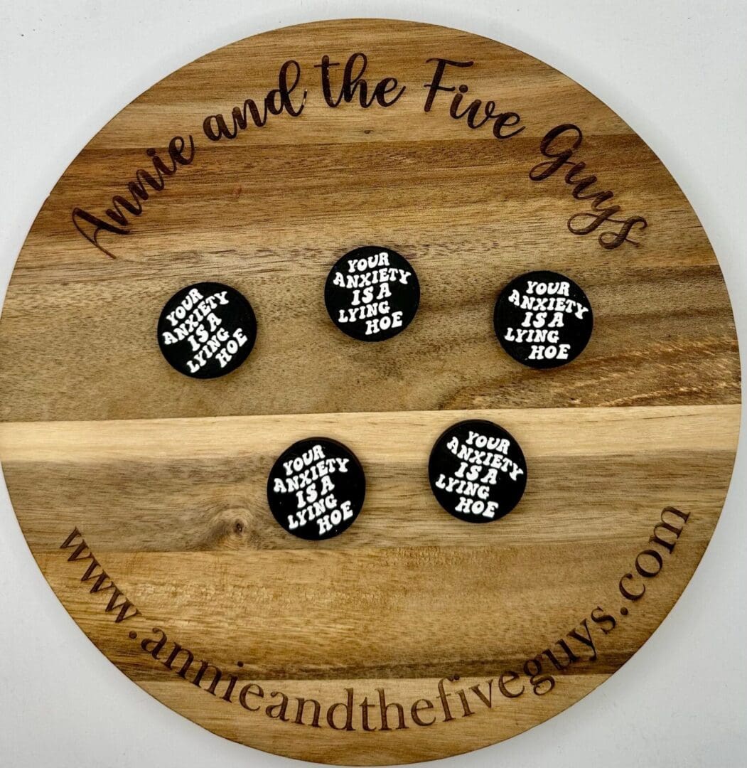 A wooden tray with "Annie and the Five Guys" engraved around the edge displays five round badges that read "Your anxiety is a lying hoe." The website "www.annieandthefiveguys.com" is also engraved.