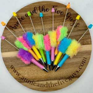 A set of neon, fluffy, colorful, plastic pens is arranged on a wooden platter with "Annie and the Five Guys" written on it and a website link below.