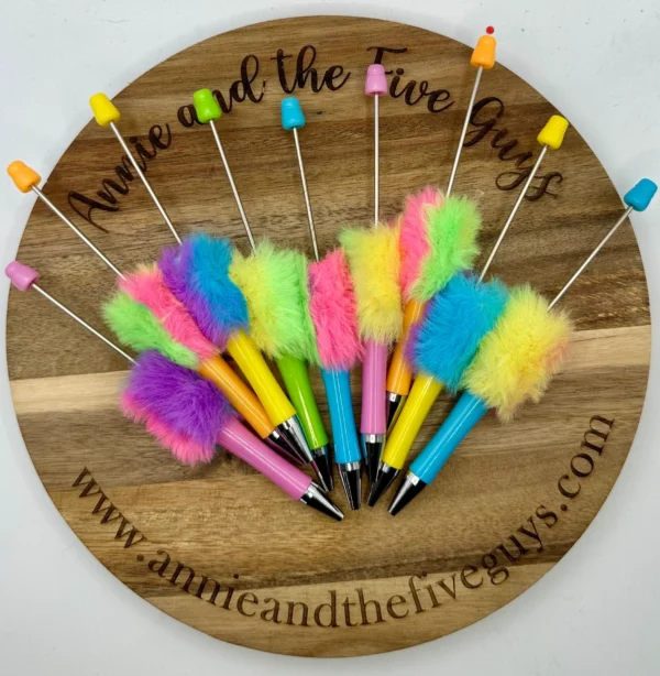 A set of neon, fluffy, colorful, plastic pens is arranged on a wooden platter with "Annie and the Five Guys" written on it and a website link below.