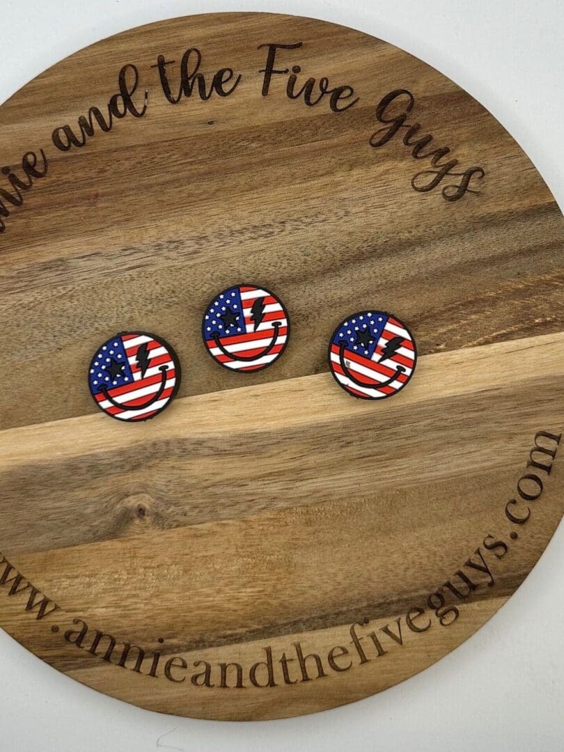 Three small, round pins with American flag designs are displayed on a wooden circular board with the text "www.annieandthefiveguys.com" around the edge.