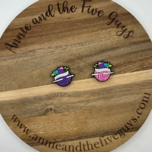 Two colorful pins shaped like yarn balls with knitting needles are placed on a wooden board with the text "Crochet all-day silicone focal bead" and a website URL around the edge.