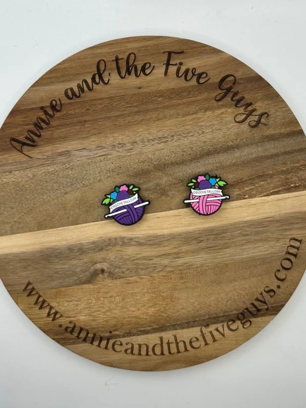 Two colorful pins shaped like yarn balls with knitting needles are placed on a wooden board with the text "Crochet all-day silicone focal bead" and a website URL around the edge.