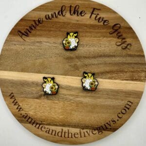 A wooden circular board engraved with "Annie and the Five Guys" and website details, displays three bee-themed pins holding small planters.