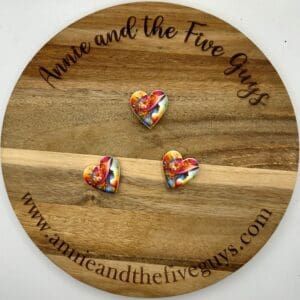 Three colorful heart-shaped pins are displayed on a round wooden plaque with the text "Annie and the Five Guys" and a website URL, annieandthefiveguys.com.