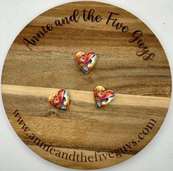 Three colorful heart-shaped pins are displayed on a round wooden plaque with the text "Annie and the Five Guys" and a website URL, annieandthefiveguys.com.