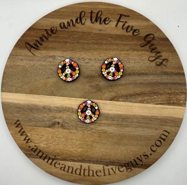 Round wooden board with the text "Annie and the Five Guys," displaying three colorful peace sign buttons decorated with floral patterns. Website "www.annieandthefiveguys.com" is also visible.