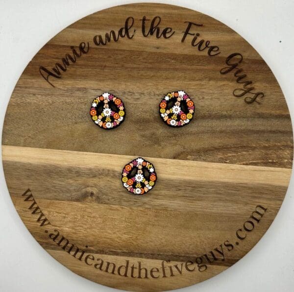 Round wooden board with the text "Annie and the Five Guys," displaying three colorful peace sign buttons decorated with floral patterns. Website "www.annieandthefiveguys.com" is also visible.