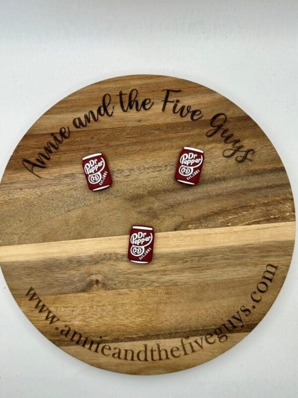 Round wooden board with the text "Annie and the Five Guys" and "www.annieandthefiveguys.com" engraved on it. Three small Dr pep silicone focal beads are placed on the board.