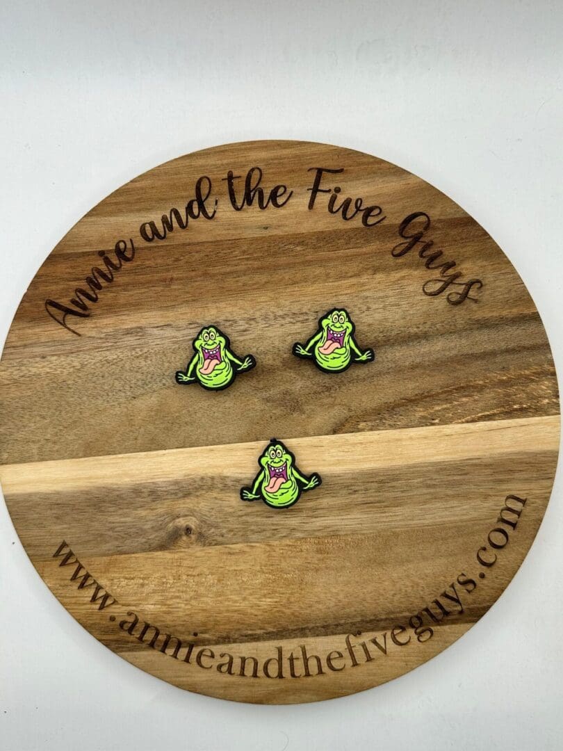 A round wooden board with the text "Annie and the Five Guys" and the website "www.annieandthefiveguys.com" displays three identical smiling cartoon character pins.