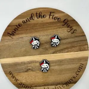 A wooden circular board with the text "Annie and the Five Guys" holds three small, pirate-themed pins featuring a black and white figure with a red bow. Website link: www.annieandthefiveguys.com.