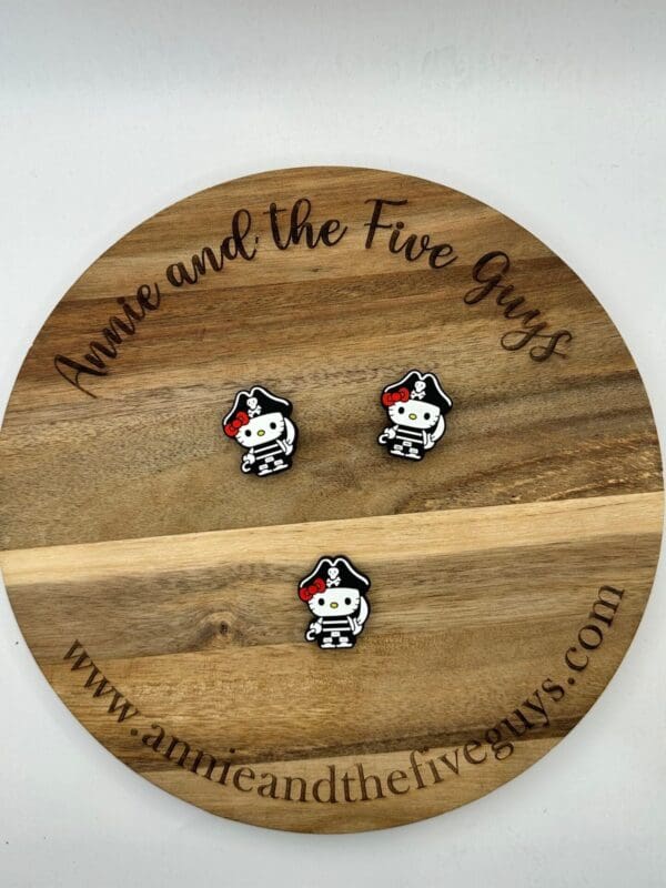 A wooden circular board with the text "Annie and the Five Guys" holds three small, pirate-themed pins featuring a black and white figure with a red bow. Website link: www.annieandthefiveguys.com.