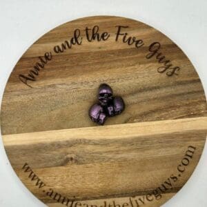 A round wooden board with "Annie and the Five Guys" and a website URL inscribed around the edges. In the center, there is a small purple metallic figure of a knight in armor.