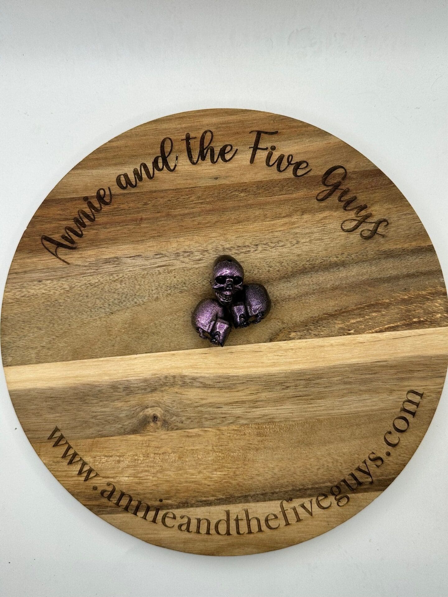 A round wooden board with "Annie and the Five Guys" and a website URL inscribed around the edges. In the center, there is a small purple metallic figure of a knight in armor.