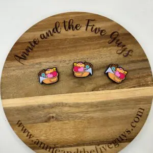 Three small colorful pins with a bird carrying a heart displayed on a round wooden board with the text "Annie and the Five Guys" engraved and their website address.