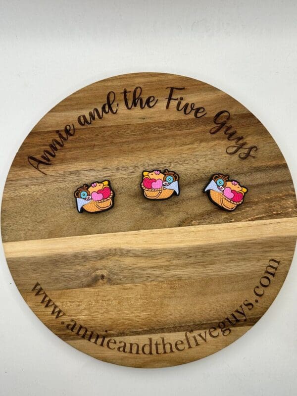 Three small colorful pins with a bird carrying a heart displayed on a round wooden board with the text "Annie and the Five Guys" engraved and their website address.